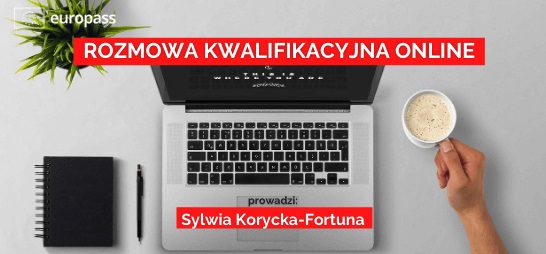 Otwarty laptop na stole, obok notes, kwiatek i ręka ludzka trzymająca kubek z kawą.