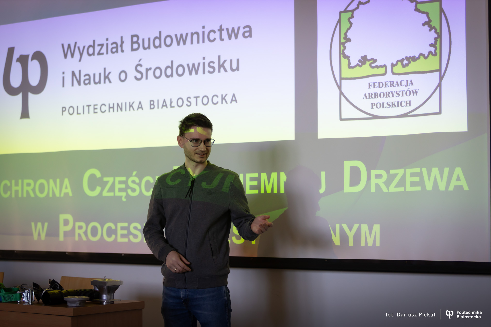 Młody mężczyzna prowadzi wykład na Wydziale Budownictwa i Nauk o Środowisku. Tło zdjęcia to prezentacja.