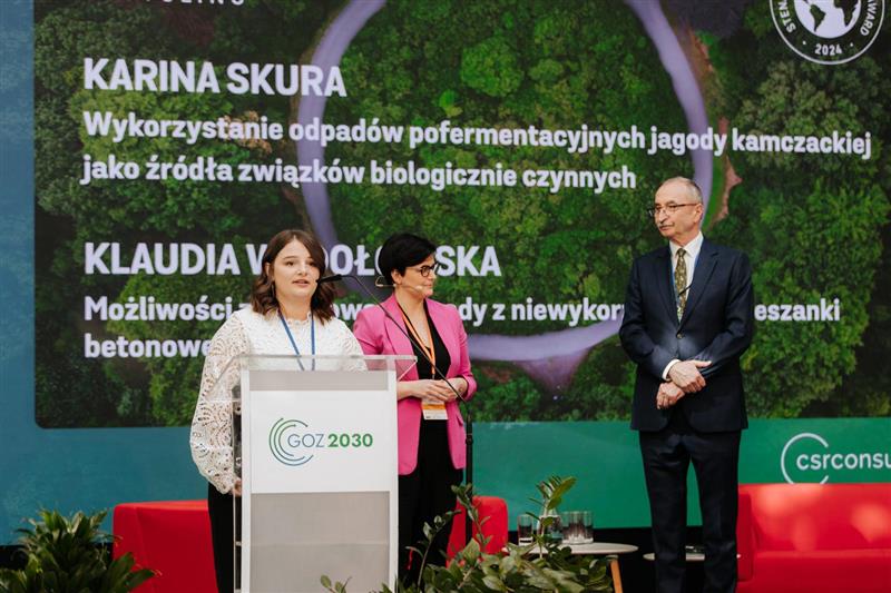 Trzy osoby elegancko ubrane, w tle różnokolorowa prezentacja. Osoba na pierwszym planie przemawia.