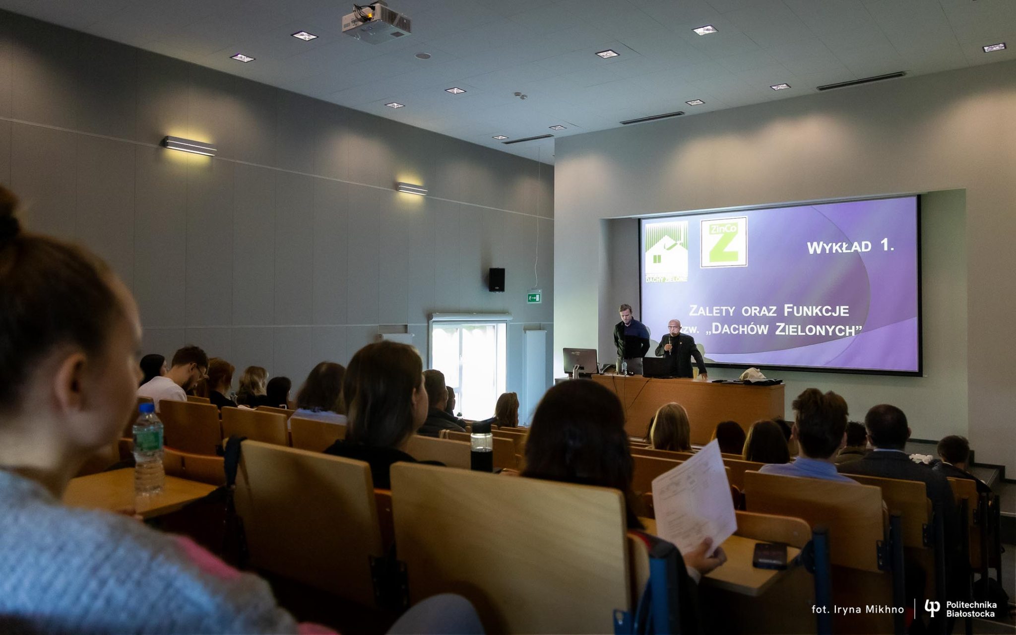 Aula wypełniona ludźmi, którzy biorą udział w szkoleniu. W tle widoczna prezentacja na rzutniku.