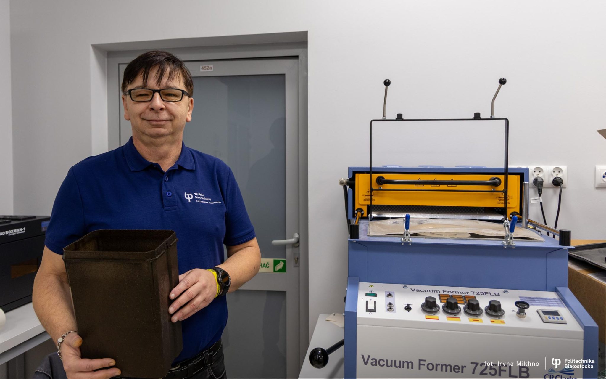 Po lewej stronie zdjęcia naukowiec w hranatowej koszulce z logiem PB, po prawej stronie maszyna do badań.