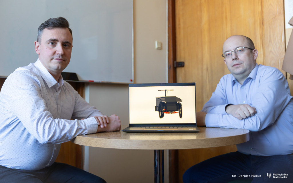 Dwie osoby siedzą przy okrągłym stole, na którym stoi otwarty laptop prezentujący wizualizację trójkołowego elektrycznego roweru cargo. W tle wnętrze pomieszczenia biurowego z tablicą i drewnianymi drzwiami.