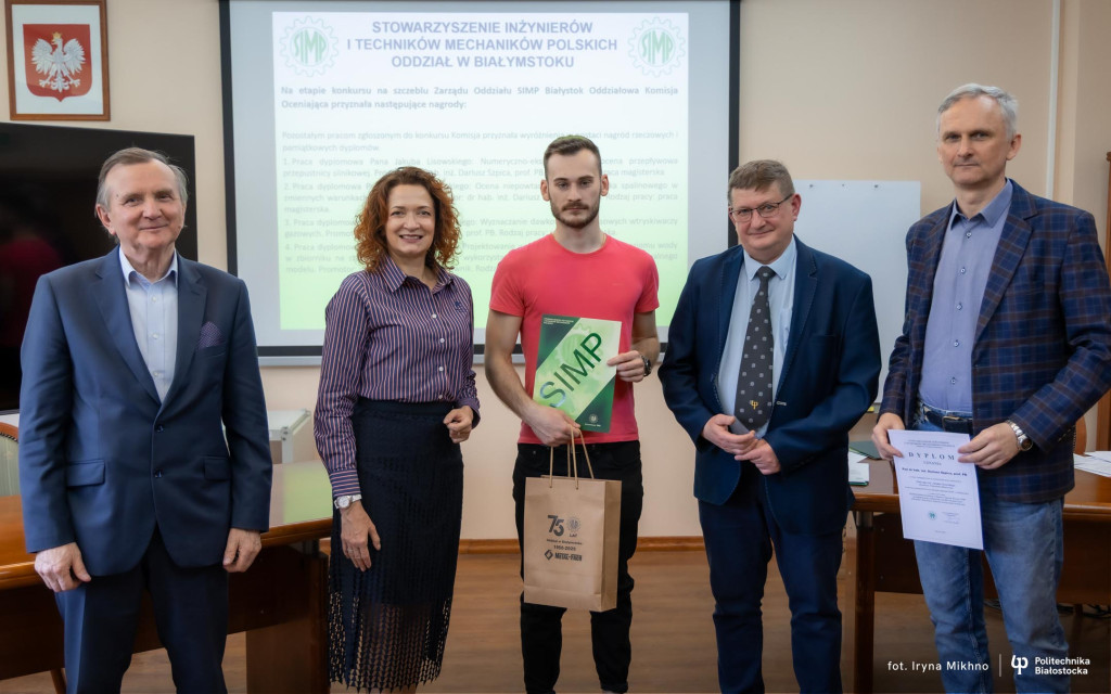 Uroczystość wręczenia nagród w konkursie na najlepsze prace dyplomowe organizowanym przez SIMP. Laureat w czerwonej koszulce stoi w otoczeniu przedstawicieli uczelni i stowarzyszenia na tle ekranu z listą nagrodzonych prac.
