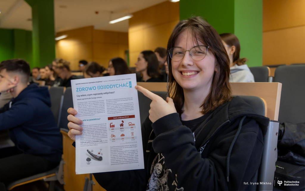 Uśmiechnięta studentka w okularach siedzi w auli wykładowej i wskazuje palcem na trzymaną w ręku ulotkę pt. "ZDROWO ((O))DDYCHAĆ". Ulotka zawiera informacje i infografiki dotyczące jakości powietrza. W tle widać innych studentów.