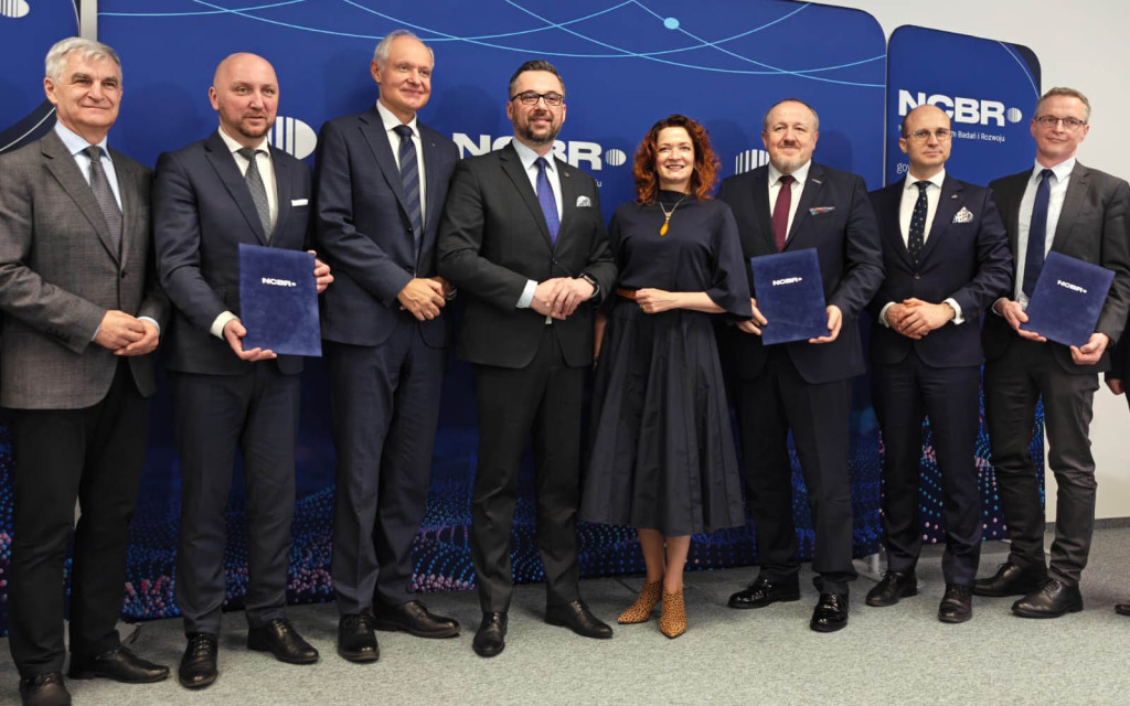 Oto propozycje tekstu alternatywnego dla tego zdjęcia, przygotowane zgodnie z zasadami dostępności: Opcja 1: Zwięzła (do mediów społecznościowych lub newsa) Oficjalne zdjęcie ośmiu osób (siedmiu mężczyzn i jedna kobieta) stojących na tle ścianki z logo NCBR (Narodowe Centrum Badań i Rozwoju). Kilku mężczyzn trzyma w rękach ciemnoniebieskie teczki z logo NCBR, co sugeruje podpisanie umowy lub wręczenie wyróżnień.