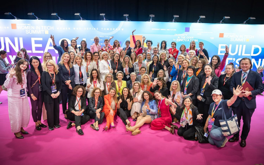 Duża grupa uczestniczek i uczestników konferencji Perspektywy Women in Tech Summit pozująca do pamiątkowego zdjęcia na jaskraworóżowym dywanie. Kobiety i mężczyźni stoją oraz siedzą na podłodze, uśmiechając się i machając w stronę aparatu na tle niebieskiej ścianki z napisami.