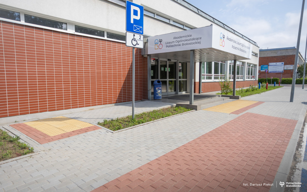 Fotografia przedstawia wejście do budynku Akademickiego Liceum Ogólnokształcącego Politechniki Białostockiej. Główne elementy zdjęcia: Wejście główne: Przeszklone drzwi wejściowe osłonięte białym zadaszeniem, na którym umieszczono logo i nazwę szkoły („Akademickie Liceum Ogólnokształcące Politechniki Białostockiej”). Infrastruktura i udogodnienia: Przed wejściem znajduje się chodnik z wyznaczonymi płytkami dotykowymi dla osób niedowidzących oraz znak drogowy informujący o miejscu parkingowym dla osób z niepełnosprawnościami. Elewacja: Po lewej stronie widoczna jest ściana budynku wyłożona rudobrązowymi płytkami, a po prawej duże okna oraz fragment zieleni. Prawy dolny róg: Podpis fotografa („fot. Dariusz Piekut”) oraz logo Politechniki Białostockiej.