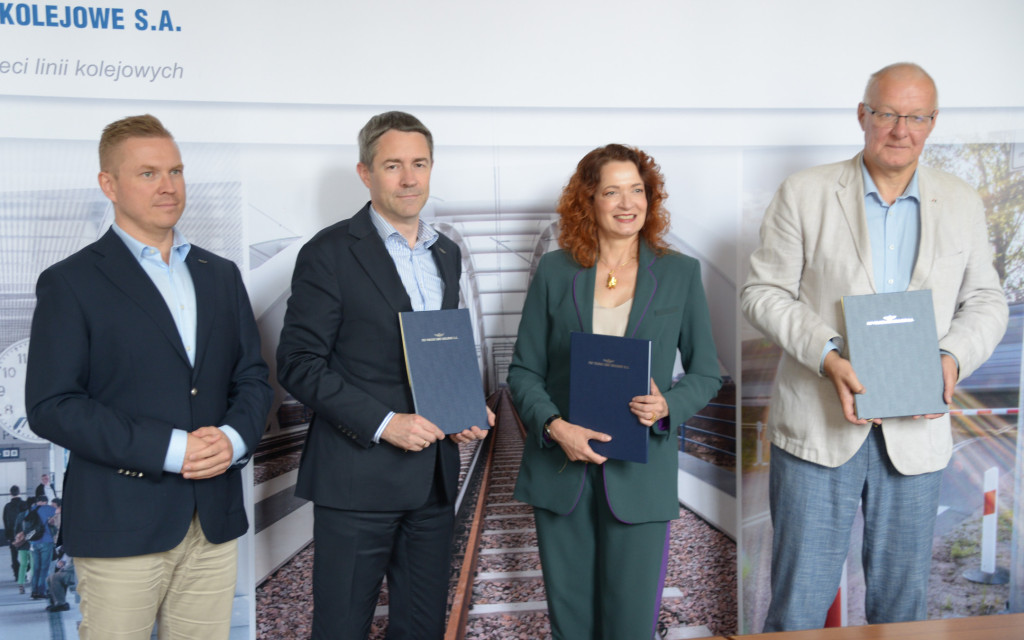 Oficjalne zdjęcie z podpisania umowy lub porozumienia. Cztery osoby — trzech mężczyzn i kobieta — stoją w rzędzie na tle ścianki z grafiką przedstawiającą tory kolejowe oraz napisem PKP Polskie Linie Kolejowe S.A. Trzy osoby po prawej stronie trzymają w rękach eleganckie, ciemne teczki z dokumentami.