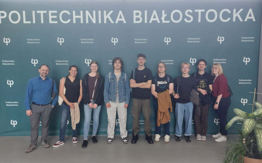Grupa dziesięciu osób stoi w jednym rzędzie na tle zielonej ścianki z powtarzającym się logo oraz dużym napisem „Politechnika Białostocka” u góry. Uczestnicy ustawieni są frontem do aparatu w holu lub przestrzeni uczelnianej. Na podłodze widoczna jest jasna posadzka, a po prawej stronie fragment rośliny doniczkowej. Zdjęcie ma charakter pamiątkowy i zostało wykonane w jasnym, wewnętrznym pomieszczeniu.