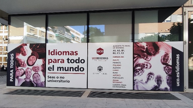 Duże okno lub przeszklona witryna budynku pokryta grafiką reklamową szkoły językowej. Na środku widoczny jest napis „Idiomas para todo el mundo” oraz „Seas o no universitario”. Obok znajduje się logo „UCO Idiomas” i lista oferowanych języków, m.in. angielski, francuski, włoski, niemiecki, chiński i arabski. Po bokach umieszczono zdjęcia grup ludzi patrzących w dół w stronę aparatu. W tle przez szybę widać odbicia budynków i ulicy.