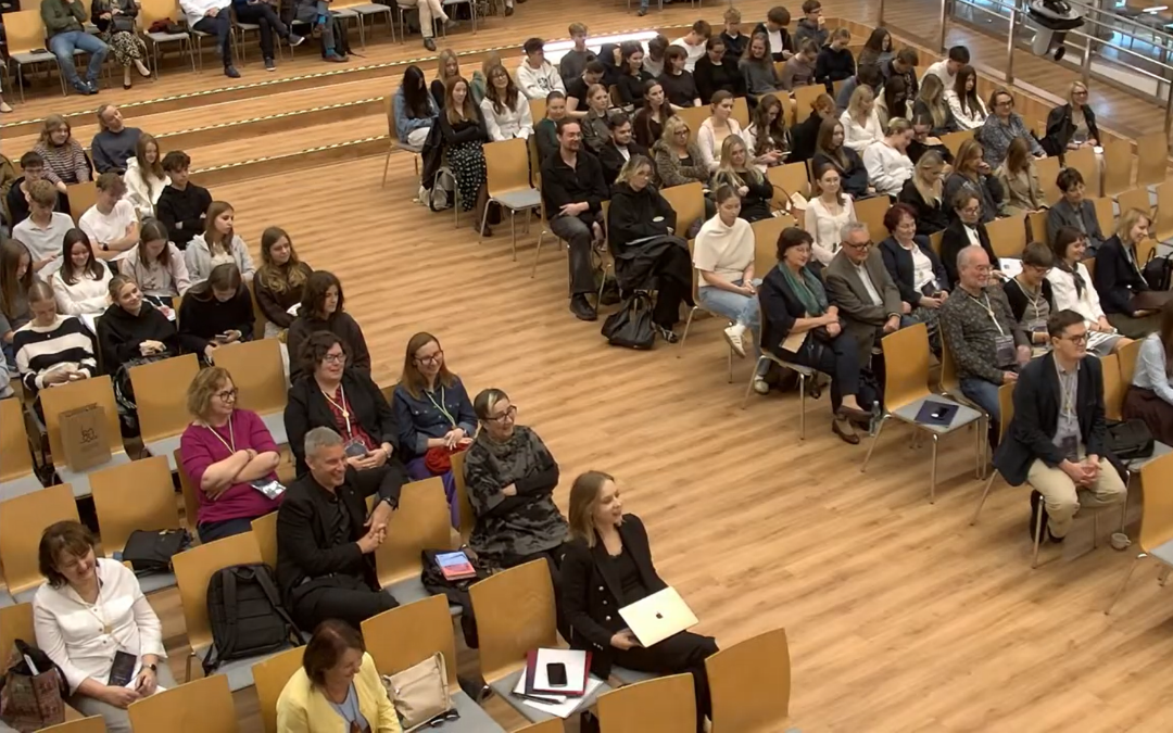 Uczestnicy konferencji lub wykładu siedzą na krzesłach ustawionych w rzędach w dużej sali audytoryjnej. W wydarzeniu bierze udział liczna grupa osób, a zdjęcie wykonano z podwyższenia, ukazując widownię oraz drewnianą podłogę sali.
