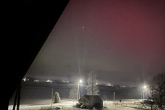Night‑time snowy landscape with a lit road, streetlights and a small building, viewed at an angle, with a pink glow in the sky