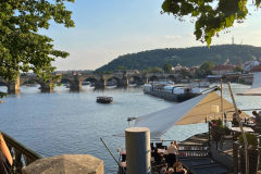 Riverfront view with a stone bridge, boats on the water and café terraces beneath leafy trees on a sunny day