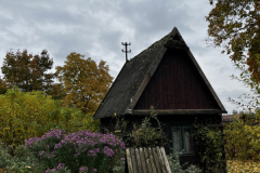 A small wooden house with a steep roof surrounded by a garden with flowers and trees with autumn foliage under a cloudy sky
