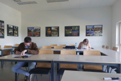 Dwie osoby siedzą przy stołach w sali i wypełniają dokumenty, na ścianie wiszą oprawione fotografie