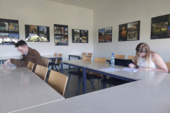 Dwie osoby siedzą przy stołach i piszą w dokumentach w jasnej sali, na ścianie widoczne są zestawy fotografii w ramkach