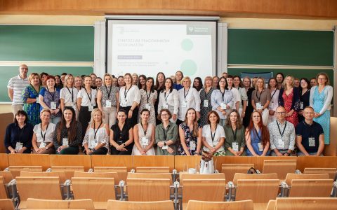 40 osób stoi na tle tablicy. 10 osób siedzi na krzesłach. Grupa jest w sali wykładowej.