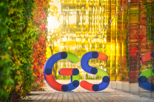 Fotografia. Duże przestrzenne i kolorowe litery GS, które są logiem portalu Geekstok ustawione na chodniku. Zdjęcie zrobione jesienią, po lewej stronie liter ściana bluszczu z liśćmi wybarwionymi na zielono i pomarańczowo, po prawej ściana szklana, w której odbija się roślinność i żółte sloneczne światlo