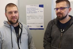 Dwóch mężczyzn pozuje na tle informacji zawieszonej na ścianie