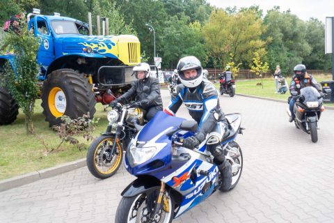 three motorcyclists on motorbikes drive through a car park, in the background on the lawn there is a blue and yellow monster truck