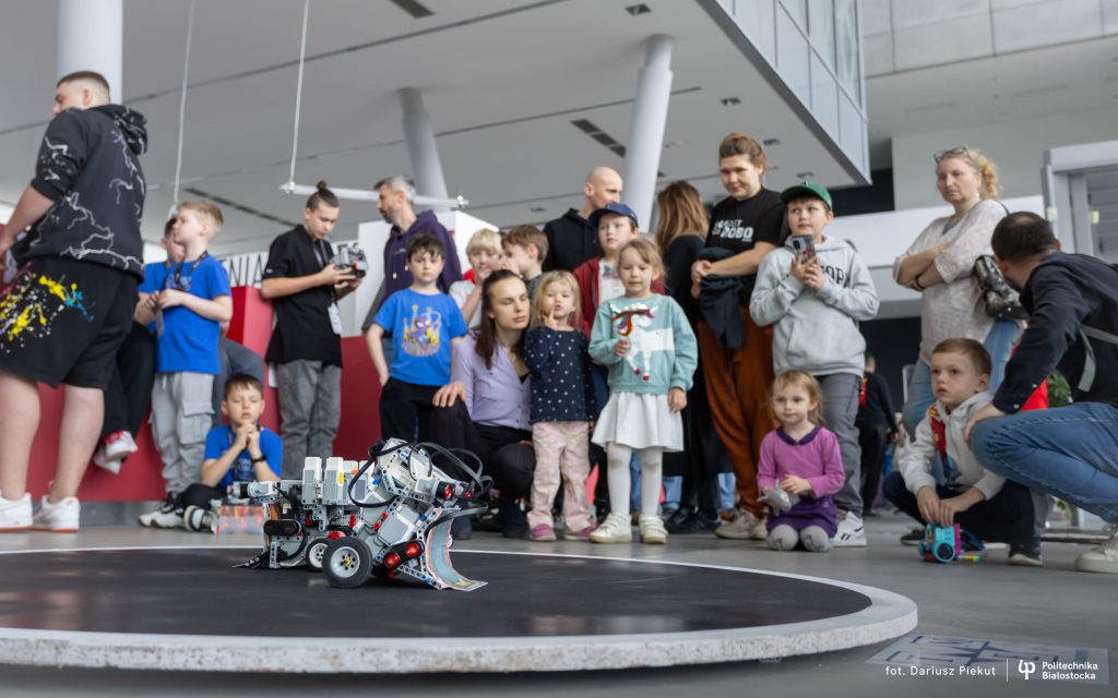 Grupa dzieci i dorosłych zgromadzona wokół okrągłej areny wewnątrz nowoczesnego budynku obserwuje małego robota mobilnego zbudowanego z klocków LEGO i elementów elektronicznych, który znajduje się na środku areny. W tle widoczne są wysokie filary i przeszklone ściany