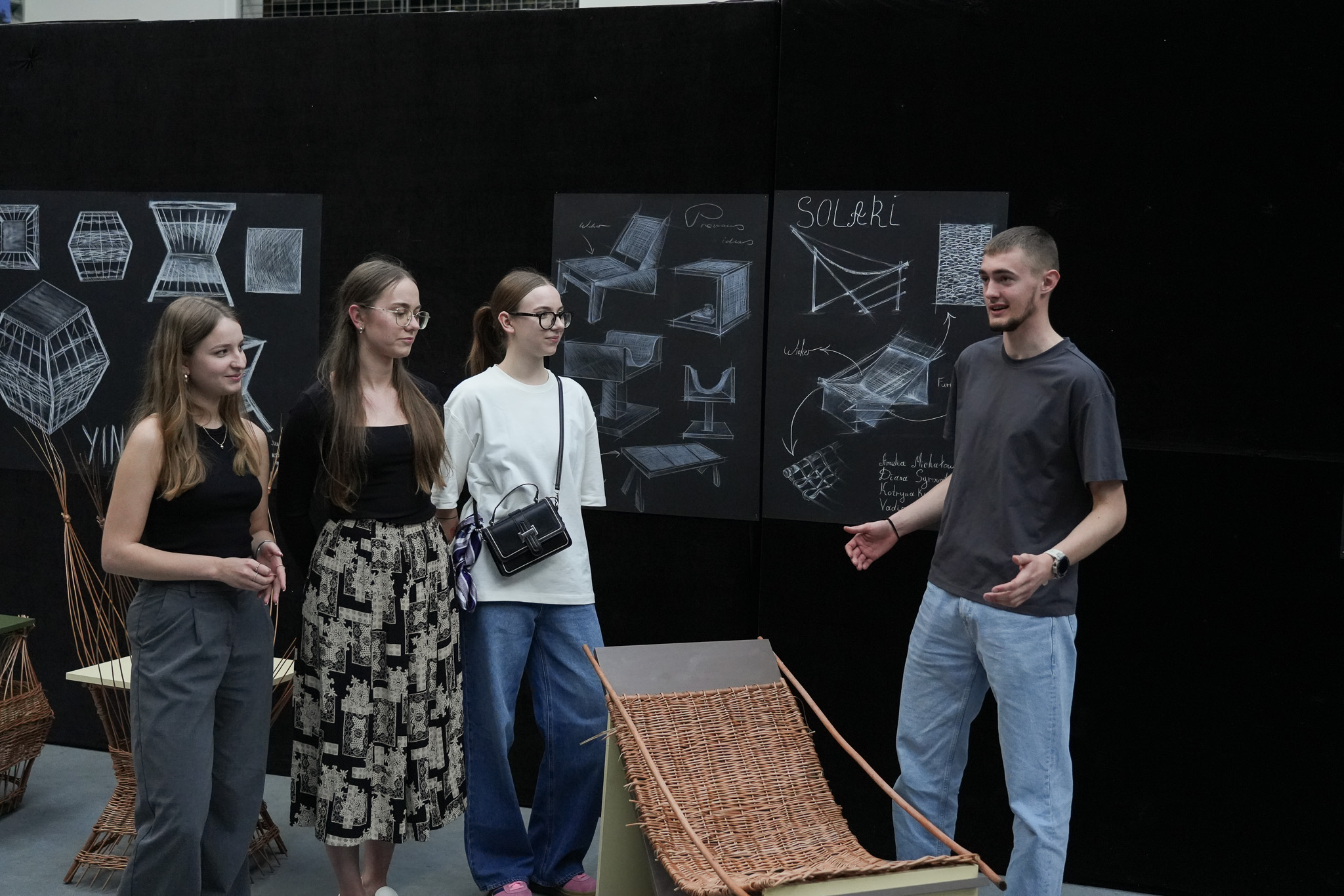 Four people are standing indoors in front of a black wall covered with boards displaying technical drawings and sketches of furniture and spatial structures. Three people are on the left, dressed in casual clothes—trousers, a skirt, and blouses. The fourth person, standing on the right, is gesturing with their hands as if explaining something to the others. In the foreground, there is an object resembling a seat or lounger, made from wicker and wood. In the background, other wicker elements are also visible.