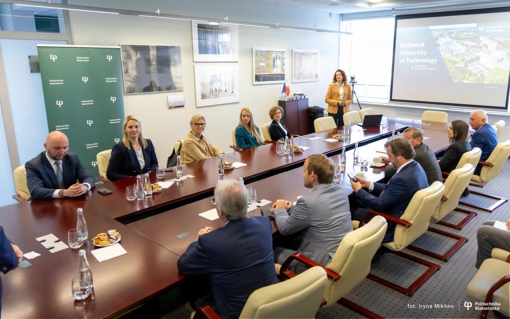 W sali konferencyjnej przy dużym, drewnianym stole siedzi kilkanaście osób, zarówno kobiety, jak i mężczyźni, w eleganckich strojach biznesowych. Na stole stoją szklane butelki z wodą, szklanki oraz talerzyki z przekąskami. W tle, po lewej stronie, widoczny jest zielony baner z logo Politechniki Białostockiej. Po prawej stronie sali, przy ekranie prezentacyjnym, stoi kobieta w żółtej marynarce, prowadząca prezentację. Na ekranie widnieje napis „Bialystok University of Technology” oraz zdjęcie kampusu uczelni. W pomieszczeniu znajdują się duże okna, które wpuszczają naturalne światło, a na ścianach wiszą obrazy