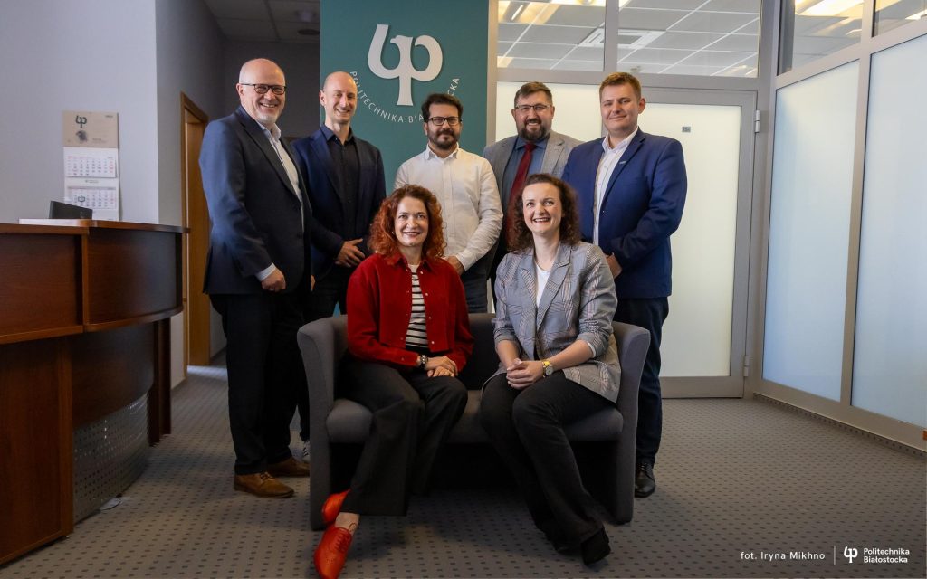 Grupa siedmiu osób, w tym kobiety i mężczyźni, pozuje do oficjalnego zdjęcia w biurowym wnętrzu. Pięć osób stoi za nowoczesną, szarą sofą, natomiast dwie kobiety siedzą na sofie z przodu. Kobieta po lewej stronie ubrana jest w czerwony żakiet, czarne spodnie i czerwone buty, a kobieta po prawej ma na sobie szarą marynarkę w kratę, białą bluzkę i czarne spodnie. Mężczyźni stojący z tyłu mają na sobie eleganckie ubrania biznesowe w odcieniach niebieskiego, szarości i bieli. W tle widoczna jest zielona ściana z logo i nazwą „Politechnika Białostocka”. Pomieszczenie posiada przeszklone ściany i drzwi, po lewej stronie znajduje się drewniana lada recepcyjna, a na ścianie wisi kalendarz. Podłoga wyłożona jest jasnoszarą wykładziną w drobny wzór.