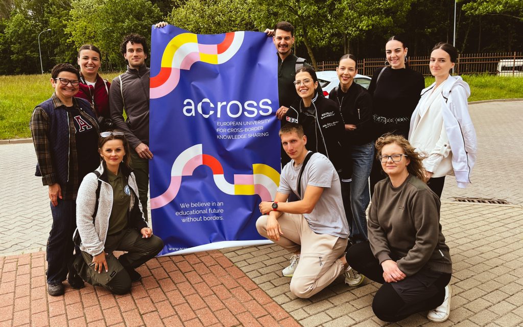 A group of people is standing and kneeling outdoors on a pavement, holding a large blue banner with the inscription: “across – European University for Cross-Border Knowledge Sharing.” The banner also features a colourful graphic with wavy lines in shades of yellow, red, pink, and white, as well as the slogan: “We believe in an educational future without borders.” In the background, there are trees, grass, a section of fencing, and a parked car. Some people are standing, while others are kneeling in front of the banner.