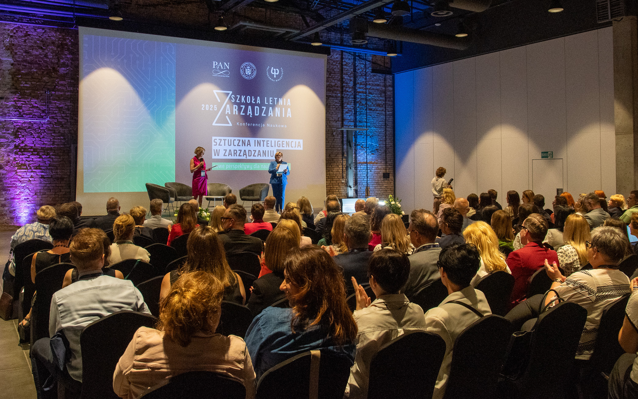 zdjęcie dużej sali konferencyjnej w której liczba publiczność siedząc przysłuchuje się dwóm prelegentkom