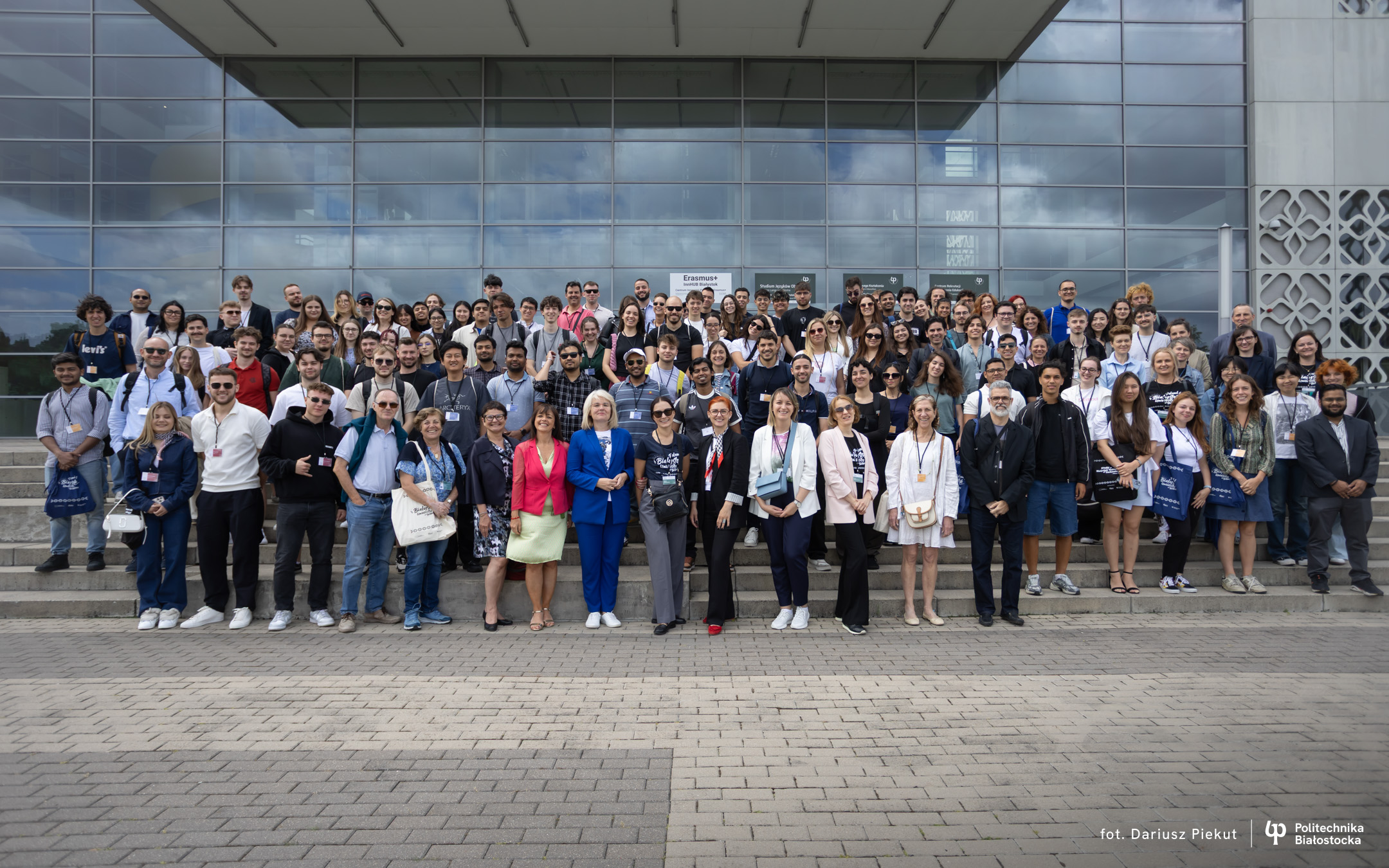 A group of around one hundred people is standing on the steps in front of a modern building with large glass windows. The participants are posing for a group photo; in the background, there is an "Erasmus+" sign