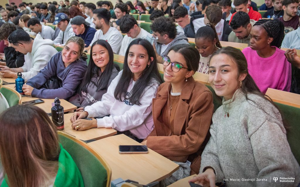 A large group of young people sit in a full lecture hall on green chairs, looking towards the speaker or talking amongst themselves; there are water bottles, phones, and notes on the desks.