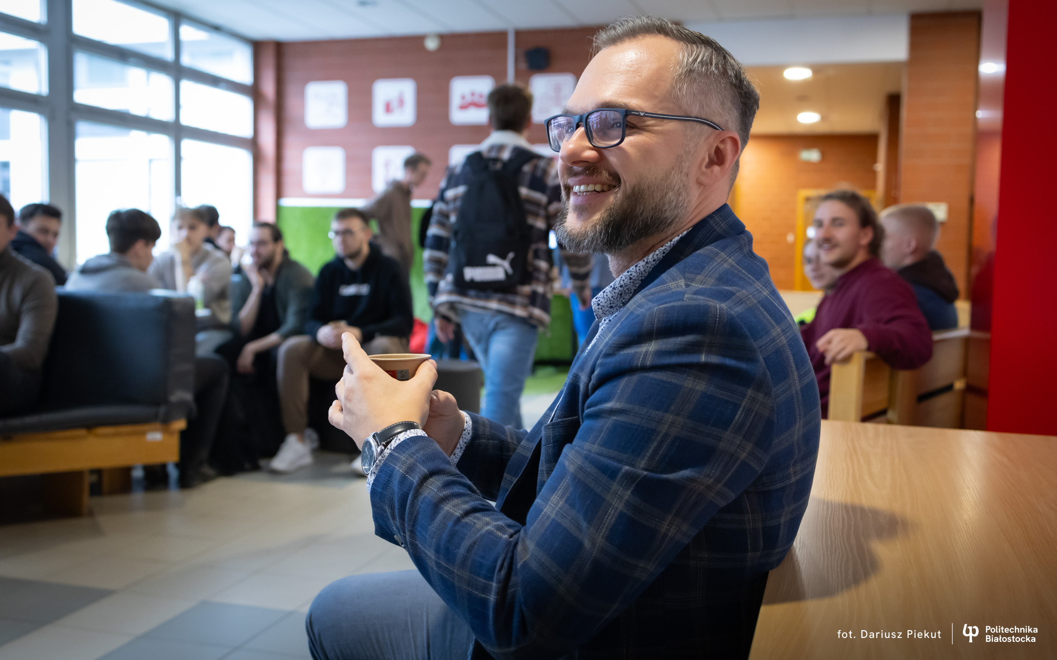 Uśmiechnięty mężczyzna, profesor Żmojda, zwrócony twarzą do studentów, w dłoniach trzyma kubek z kawą. Siedzi na drewnianej ławie, na drugim planie siedzący przy stolikach z kawą i ciastkami studenci