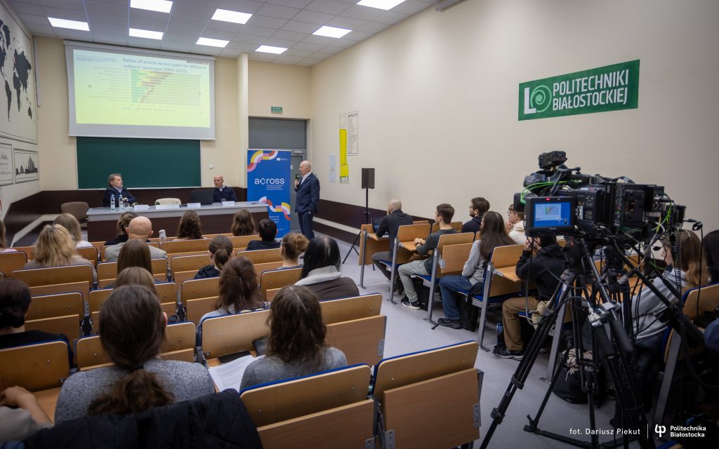 A lecture hall filled with attendees sitting on wooden benches, facing a speaker with a microphone standing next to a table with two seated men. A chart is projected on a screen at the front, a colourful banner with the word "across"