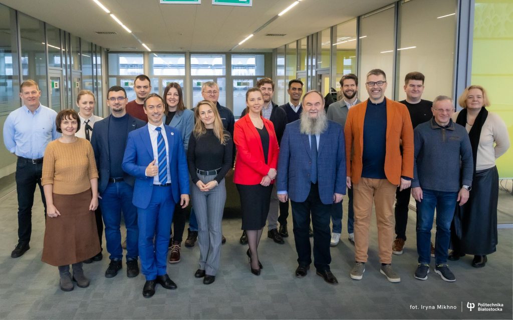 Grupa osób stoi w jasnym holu uczelni, ustawiona w kilku rzędach, pozując do wspólnego zdjęcia w przestrzeni biurowo‑konferencyjnej