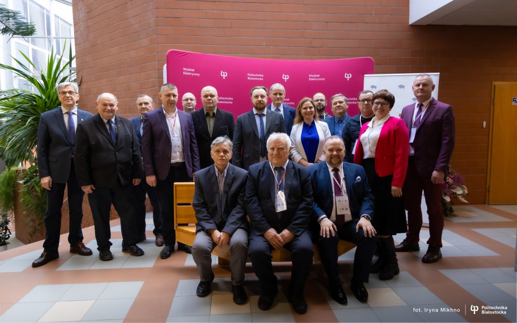 Grupa osób w formalnych strojach pozuje do wspólnego zdjęcia przed różową ścianką konferencyjną w holu budynku