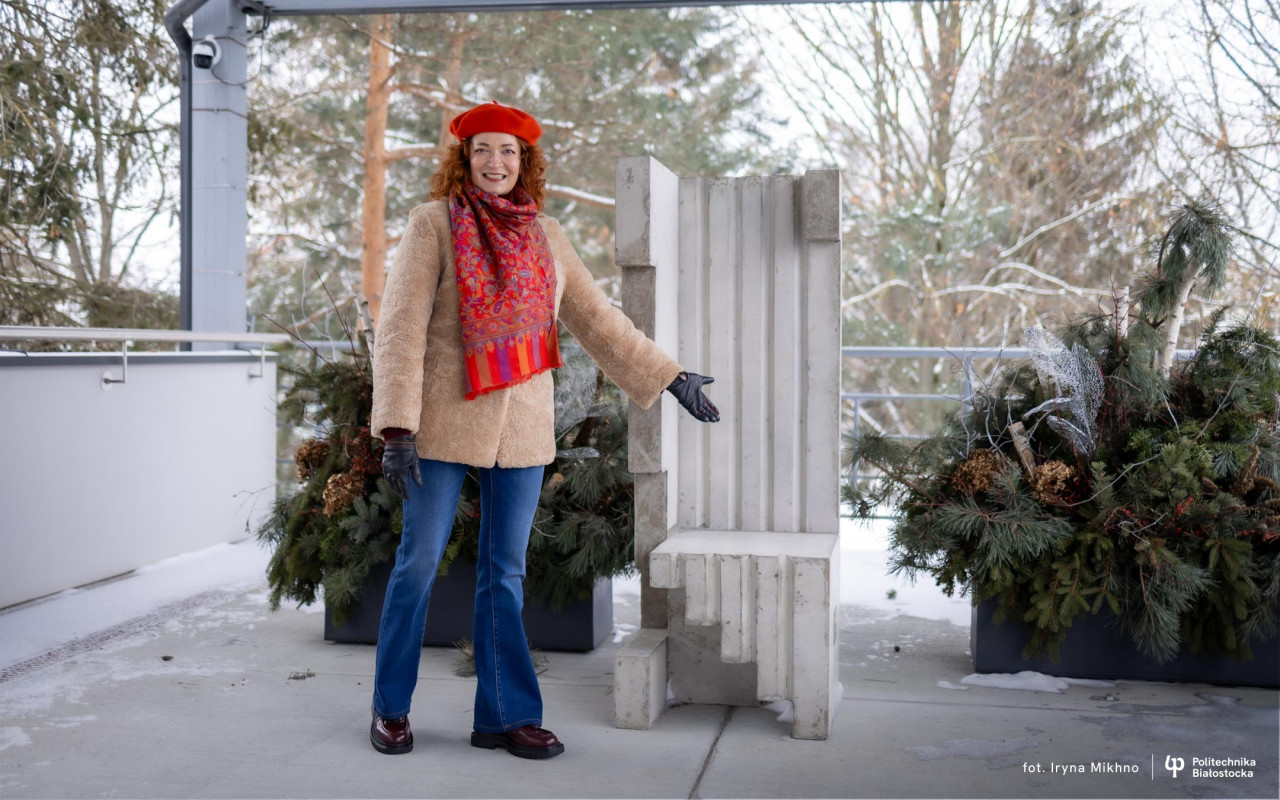 A person in winter clothing presents a concrete sculptural or architectural piece on an outdoor terrace decorated with evergreen arrangements in snowy surroundings