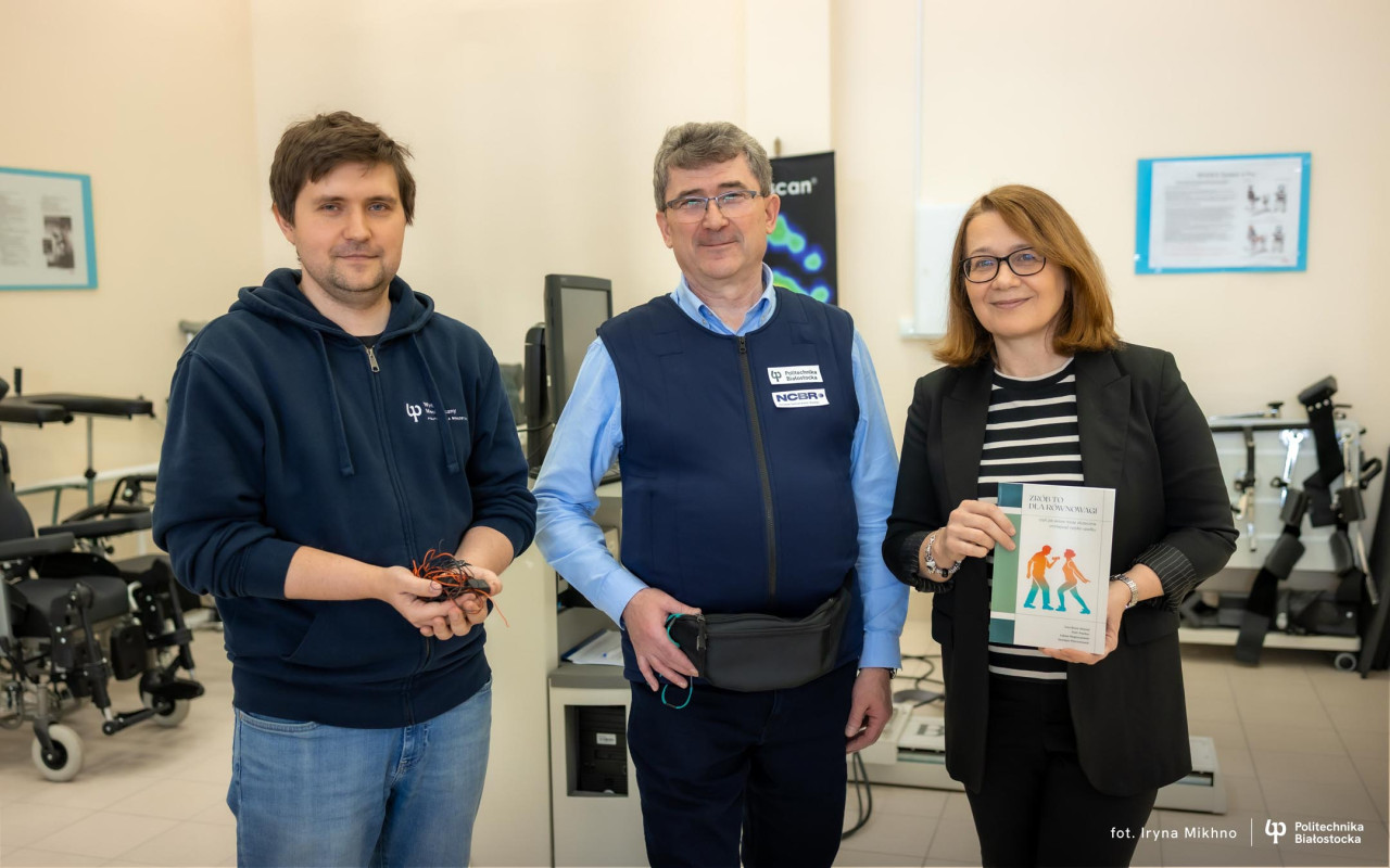 A group of people stand in a rehabilitation studio among exercise equipment, holding an electronic device and a health‑related publication