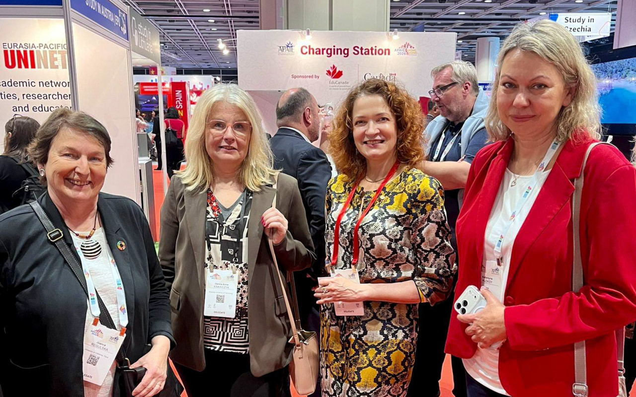 A group of people stand at an education‑industry event near a “Charging Station” booth, surrounded by banners and other participants