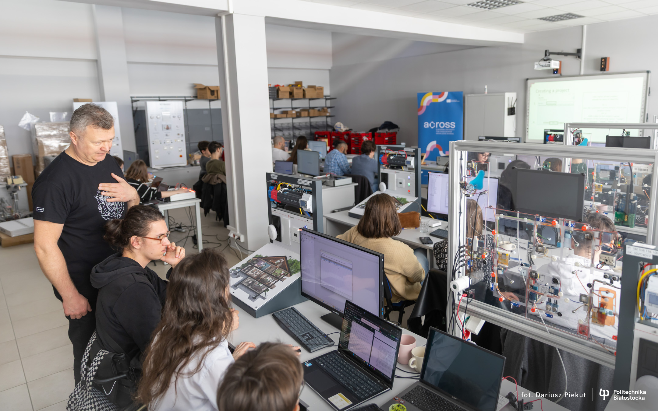 People work at computer stations in an automation laboratory surrounded by technical equipment, monitors and training apparatus