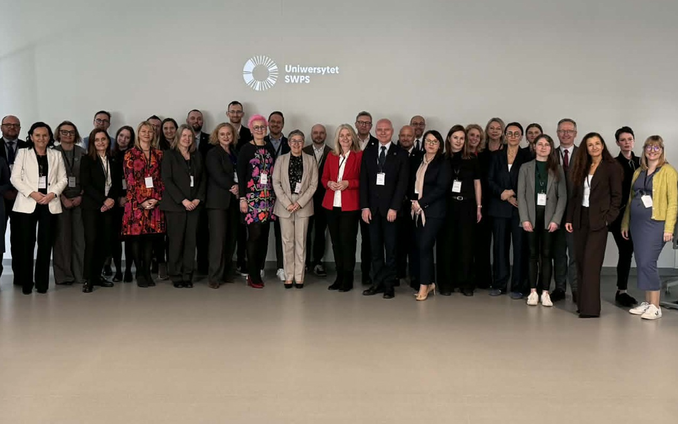 Grupa osób w eleganckich strojach stoi na tle jasnej ściany w sali konferencyjnej.