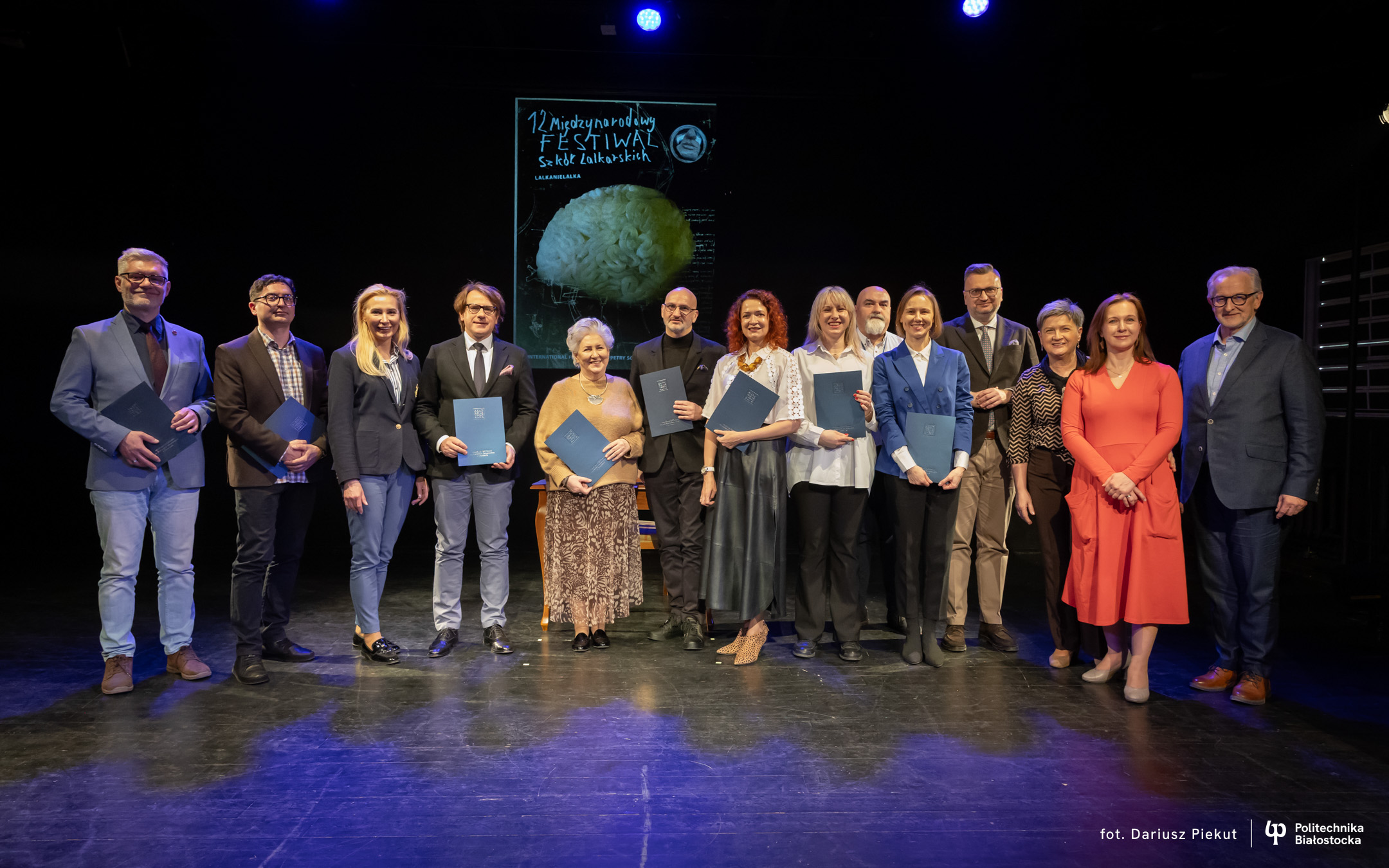 A group of people stand on a theatre stage holding certificates and posing for a group photo against an event poster backdrop