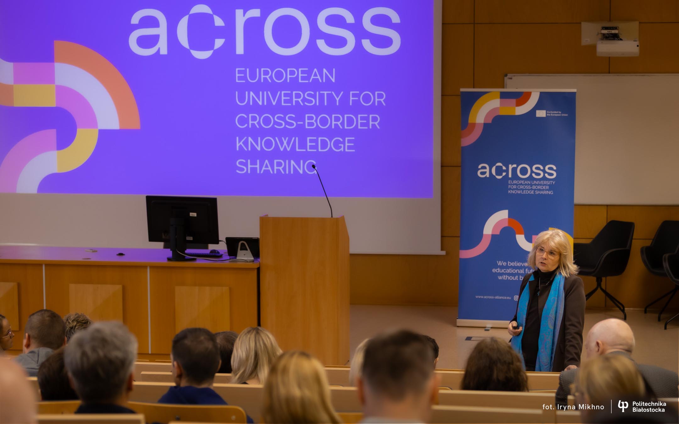 Lecture hall scene with a woman addressing seated attendees, with an Across presentation displayed in the background