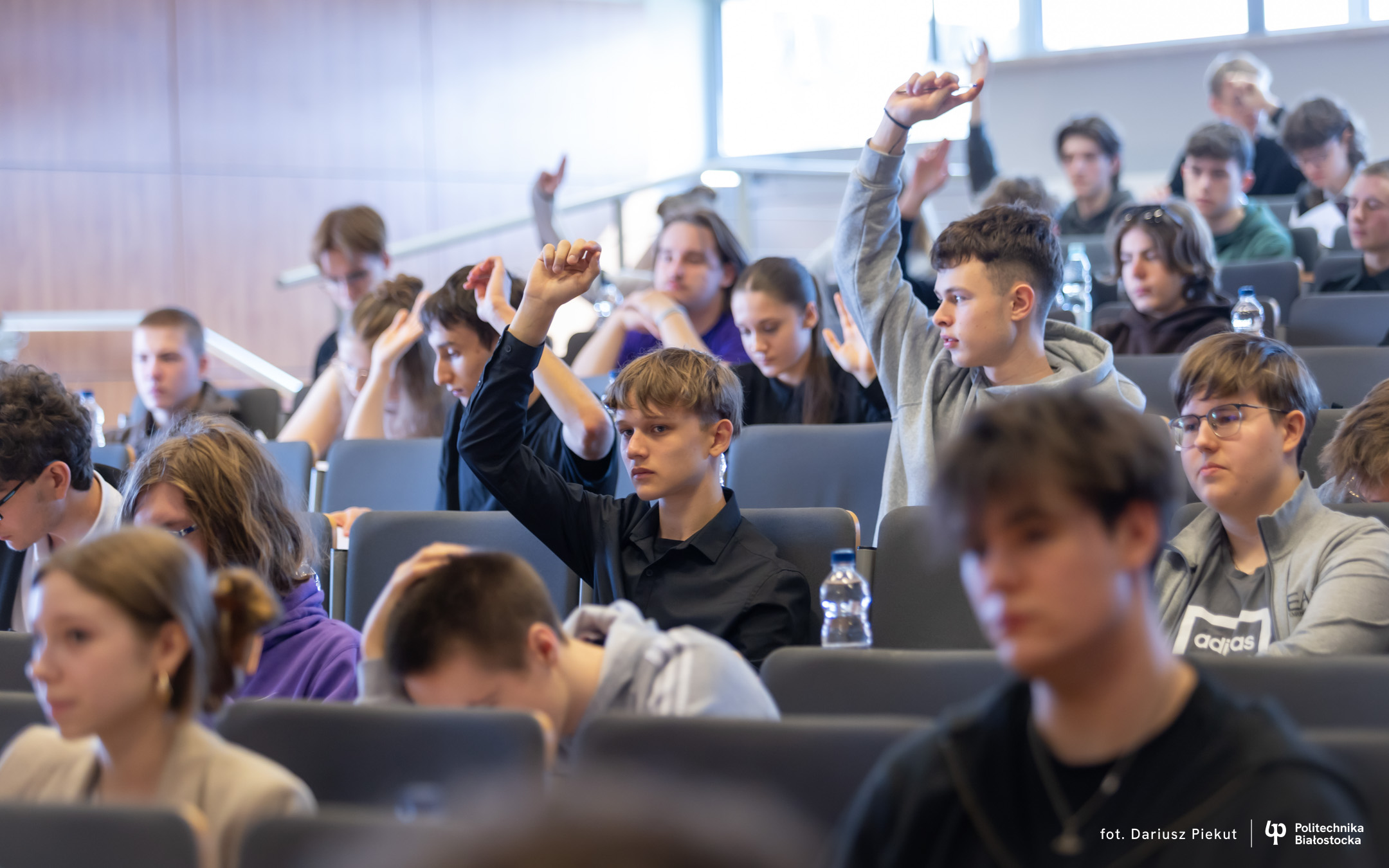 grupa uczniów siedzi na auli. podnoszą jedną rękę w górę