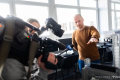 A person gives an interview in a laboratory, gesturing toward technical equipment while a camera crew records the scene